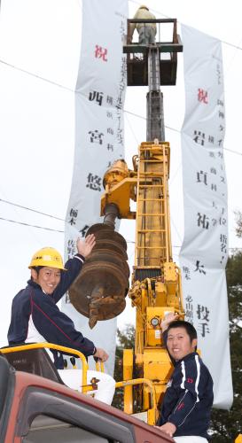 横浜商大野球部グラウンドのネットに設置された特大の垂れ幕の前で、重機に乗ってポーズを決める阪神１位指名の岩貞（左）と楽天５位指名の西宮