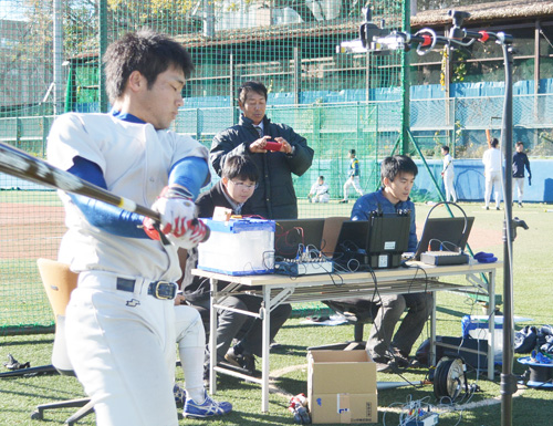 スイングスピードを計測する選手の後ろで、携帯電話のカメラで撮影する東大・浜田監督
