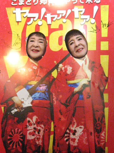 姉妹の本拠地・浅草で行われた「映画　こまどり姉妹がやって来たヤァ！ヤァ！ヤァ！」ＤＶＤ化記念イベント。普段着だからと派手な看板に顔をのぞかせるこまどり姉妹
