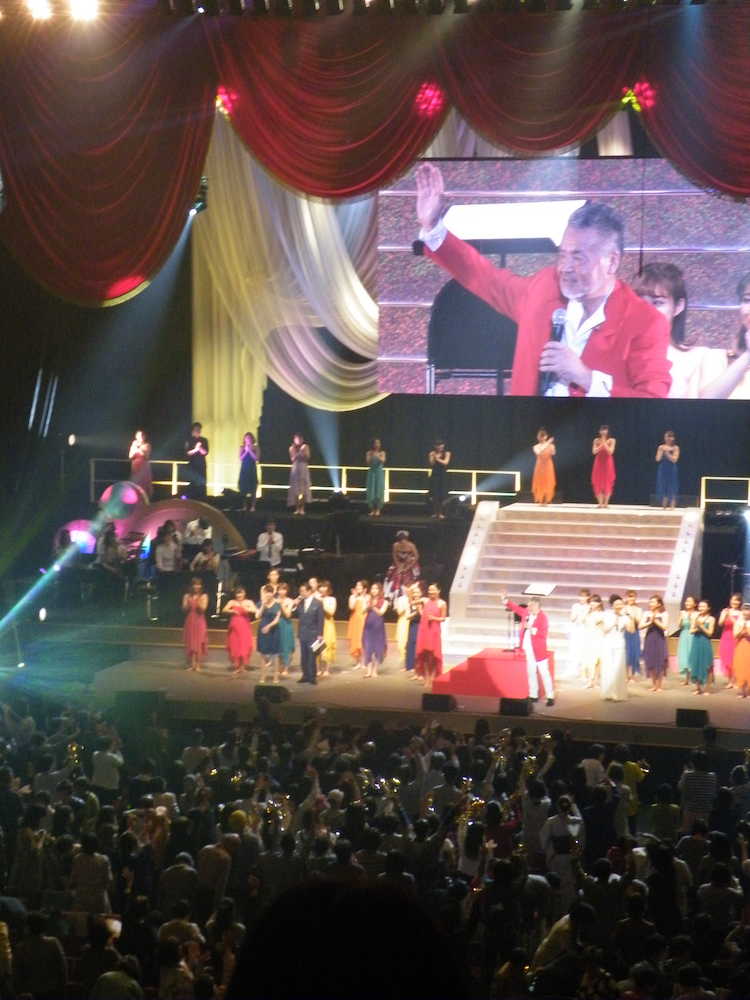 「カラオケレインボー　１万人の歌謡曲」イベントを行った中村泰士氏（階段下の赤い服、後方の映像）