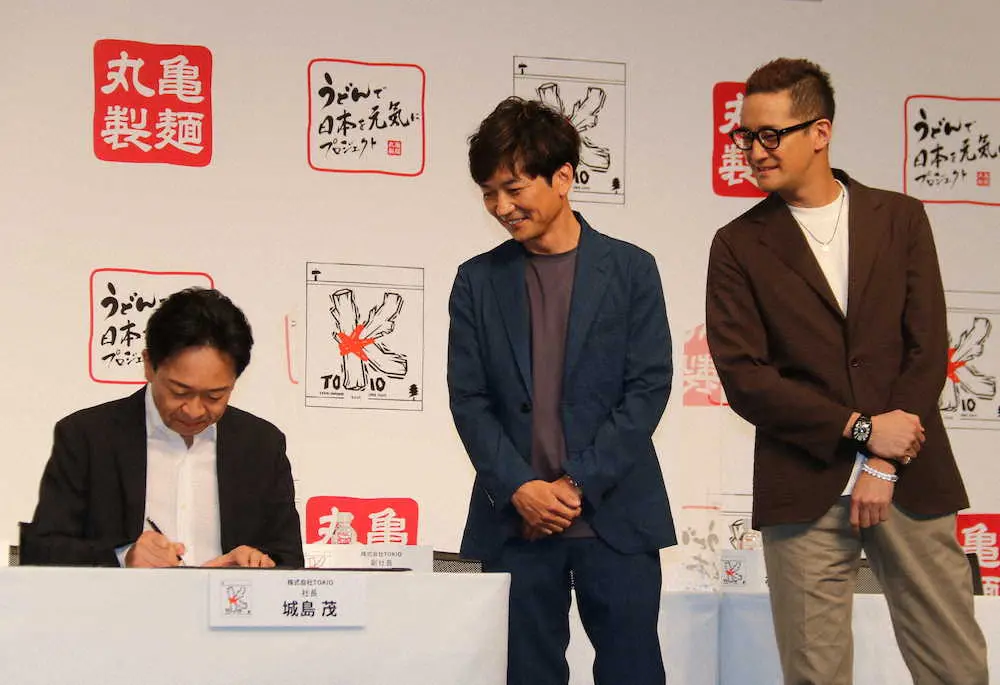 「うどんで日本を元気にプロジェクト」合同記者会見に調印式でサインする株式会社TOKIOの城嶋茂社長（左）を覗き込む、国分太一副社長（中央）、松岡昌宏副社長