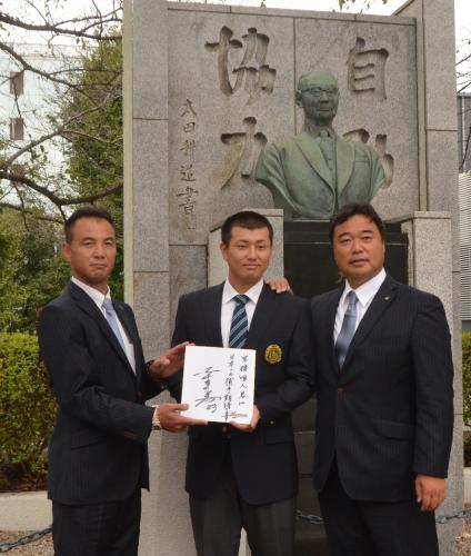 亜大の初代学長・太田耕造氏の像の前で諸積スカウト（左）から色紙を受け取る宗接。右はロッテ・永野チーフスカウト