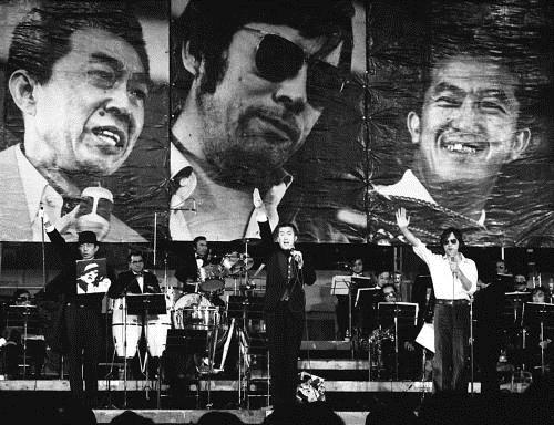 １９７４年１２月、永六輔（中央）、野坂昭如（右）と中年御三家を結成し、日本武道館でコンサートを行った小沢昭一さん。背景の写真は、左から小沢さん、野坂、永