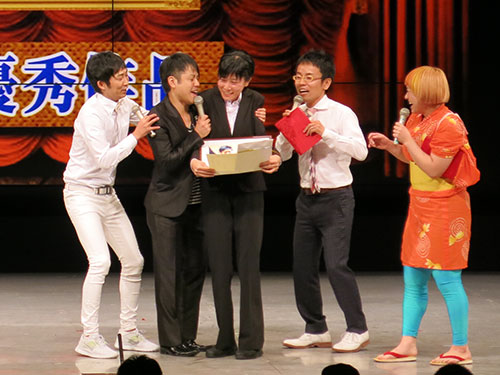 受賞女性（中央）にステージ上で抱きつく井上裕介（左から２人目）と止める相方の石田明（左）