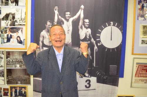 １９６４年の東京五輪重量挙げ金メダリストの三宅義信さん。当時の会場、渋谷公会堂で表彰台の中央に立った写真を背に笑顔