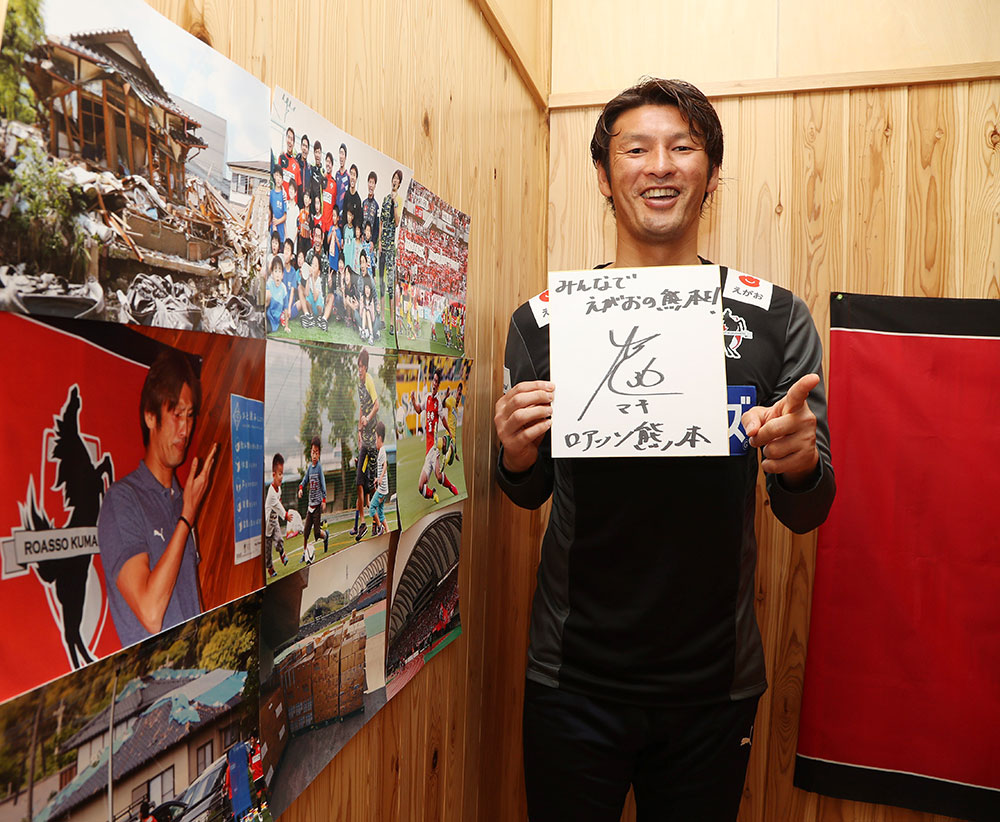 熊本地震からの１年を振り返った写真を横に、「みんなでえがおの熊本を！」と記した色紙を手にする熊本・巻