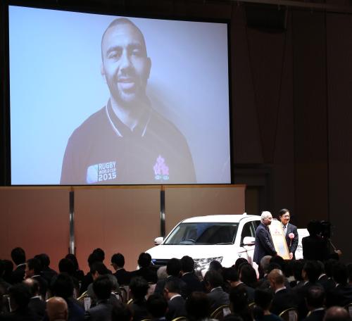 大賞を受賞し副賞の自動車の前で記念撮影する日本ラグビー協会・岡村正会長（左）。スクリーンにはリーチ主将
