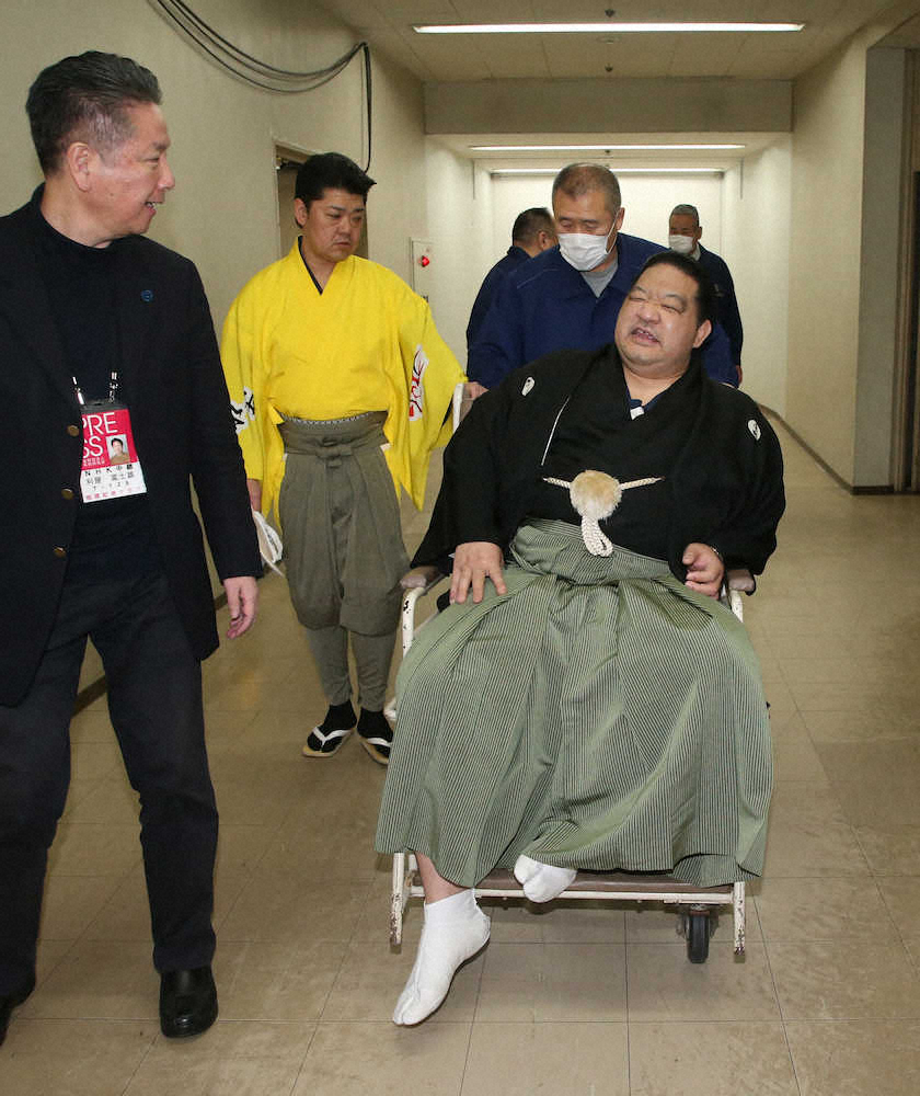 NHKの刈屋富士雄アナウンサー（左）に心配そうに見守られながら車椅子で運ばれる浅香山親方（右）（撮影・西海健太郎）